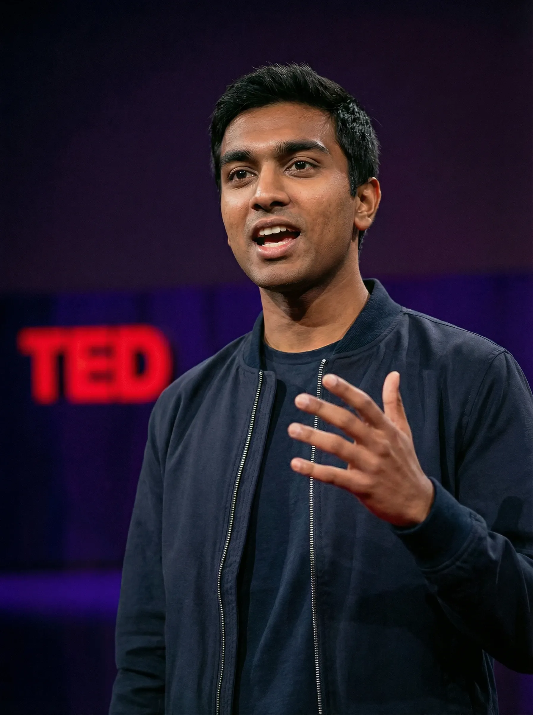 Professional speaker headshot of a man in a navy bomber jacket and dark crew neck on stage with a TED backdrop